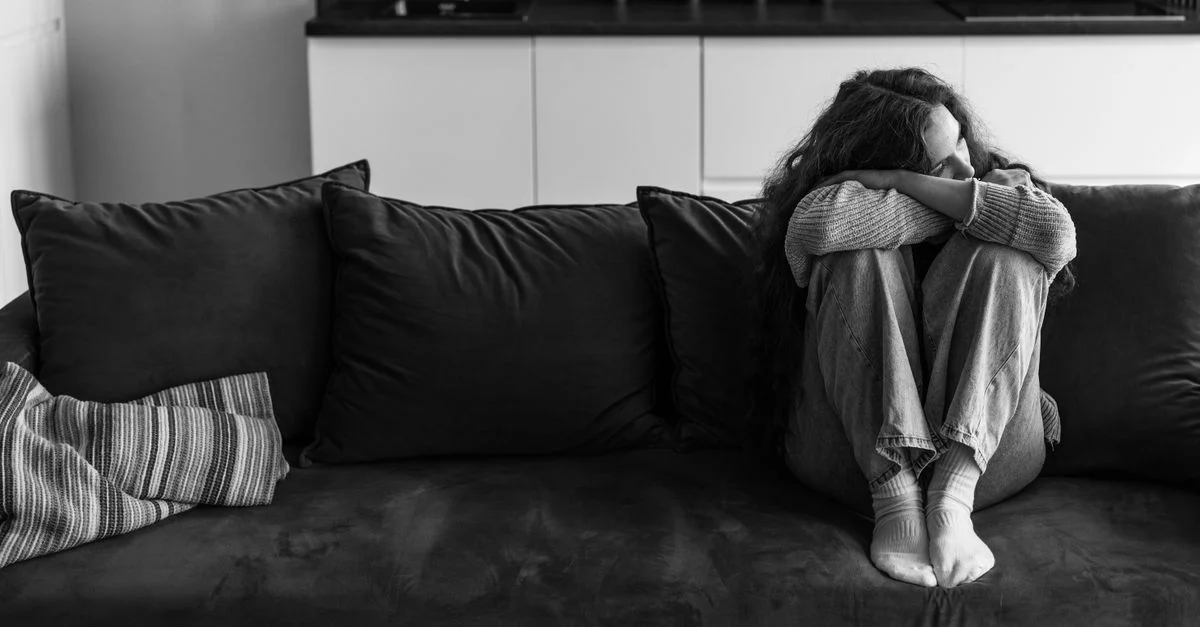 Une jeune femme est assise seule sur un sofa, affichant des émotions de tristesse et de contemplation.