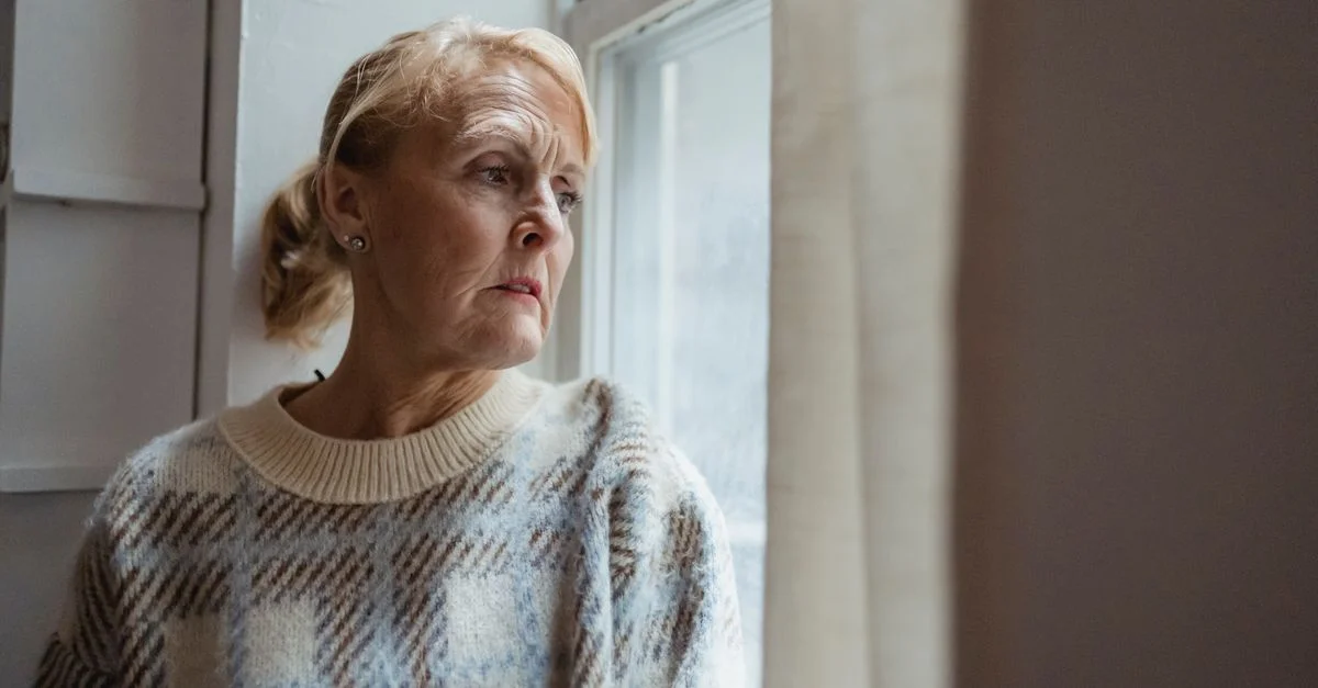 Une femme senior en profonde réflexion regardant par une fenêtre à l'intérieur.
