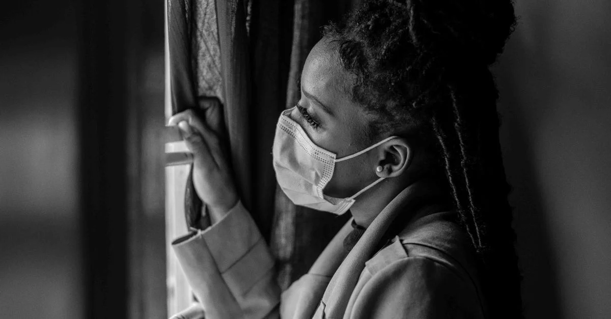 Portrait en noir et blanc d'une femme portant un masque facial, regardant par une fenêtre ouverte pendant le confinement pandémique.