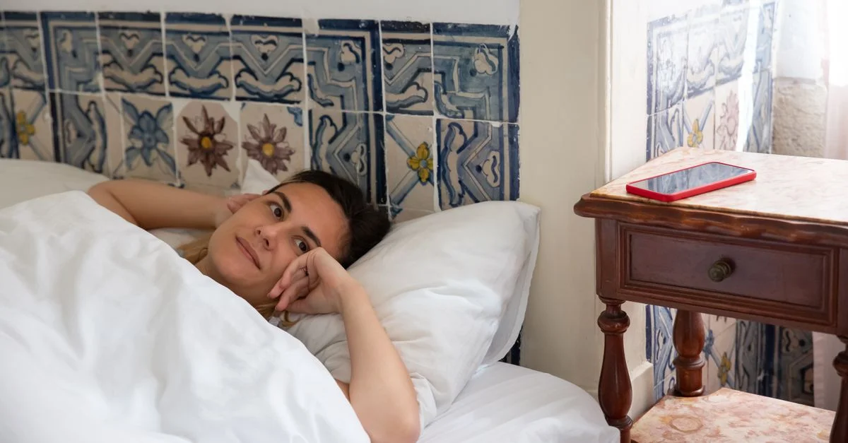 Une femme est allongée dans un lit dans une chambre carrelée au Portugal avec un smartphone sur une table de chevet.