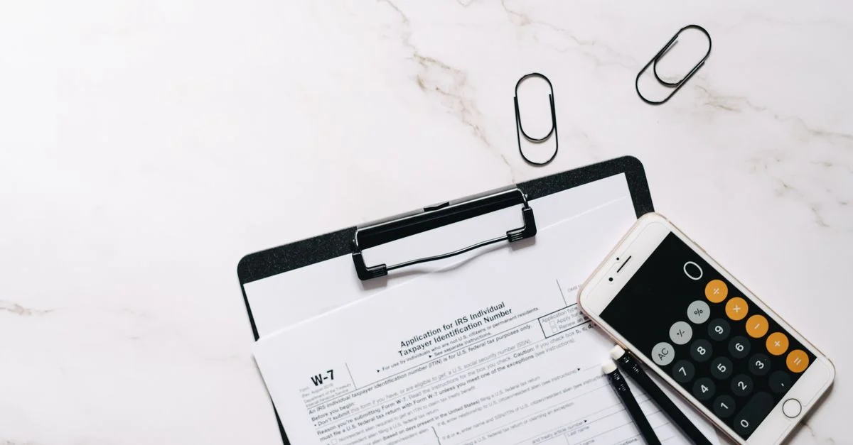 Une vue de dessus d'un formulaire W-7 de l'IRS, d'une calculatrice smartphone et de trombones pour la préparation fiscale.
