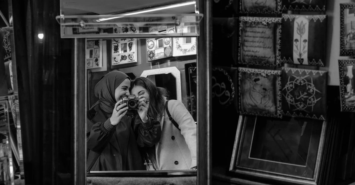 Deux femmes souriantes dans un miroir, capturant un moment avec un appareil photo dans une boutique remplie d'art.