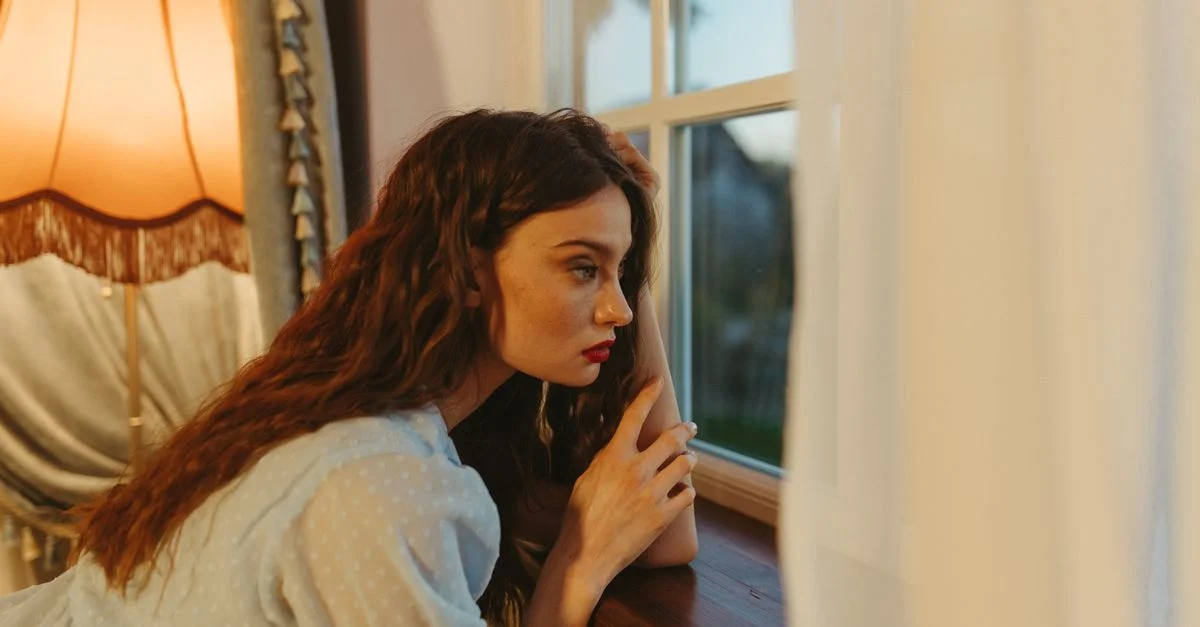 Une jeune femme aux cheveux rouges regarde pensivement par la fenêtre à l'intérieur, entourée d'une lumière chaude.