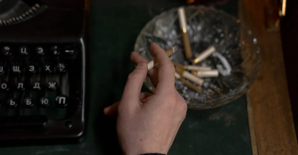 Gros plan d'une main tenant une cigarette près d'une machine à écrire vintage et d'un cendrier, évoquant l'inspiration créative.